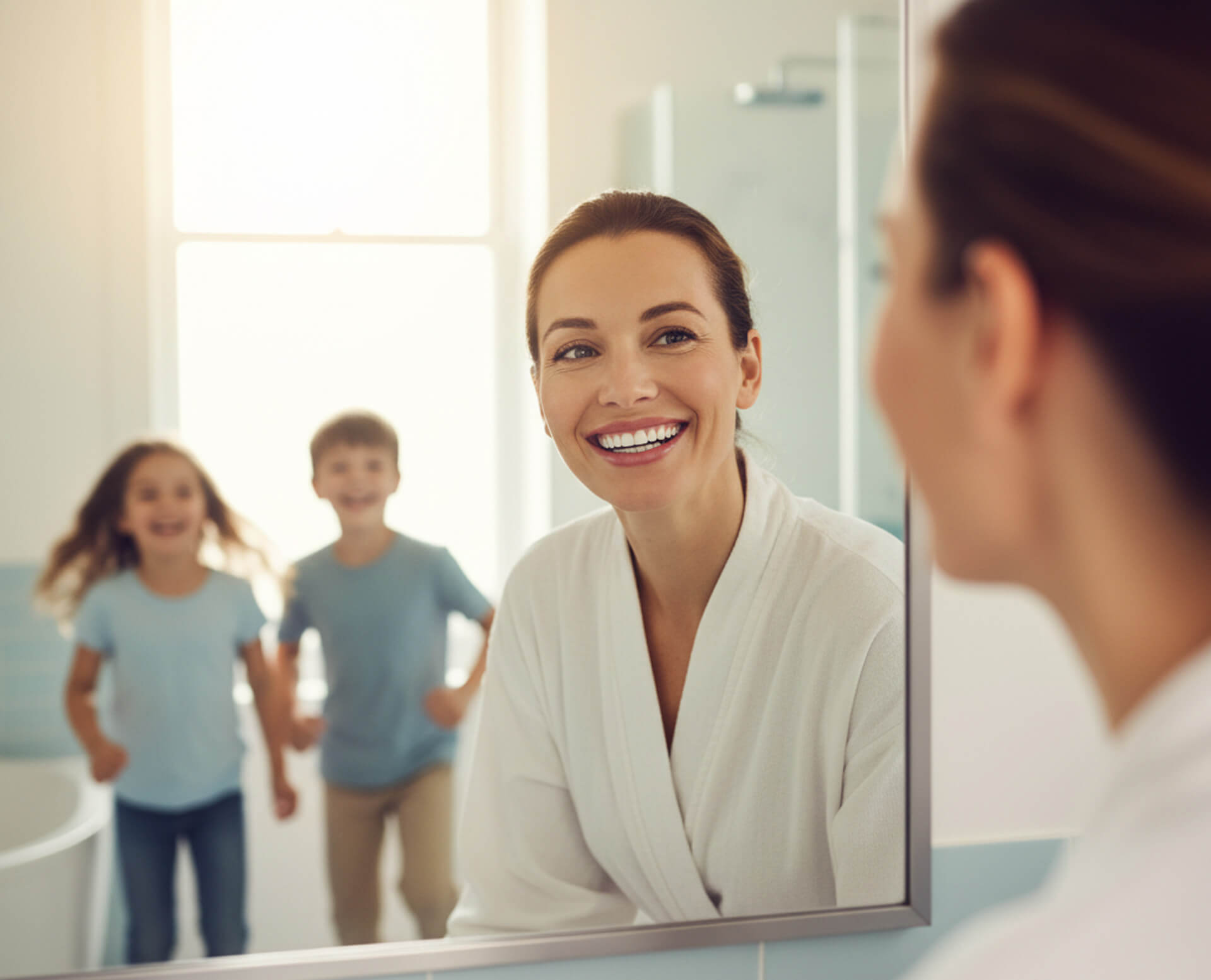 Smiling woman in a bathroom mirror, showcasing a bright smile, with two children playfully running in the background, emphasizing family joy and confidence.