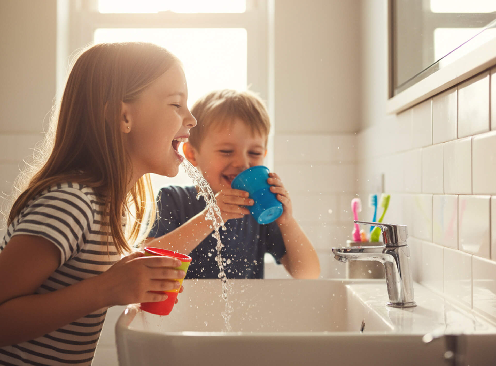 Children playing with water at a sink, one girl with a red cup and a boy with a blue cup, emphasizing fun and education in dental hygiene practices.