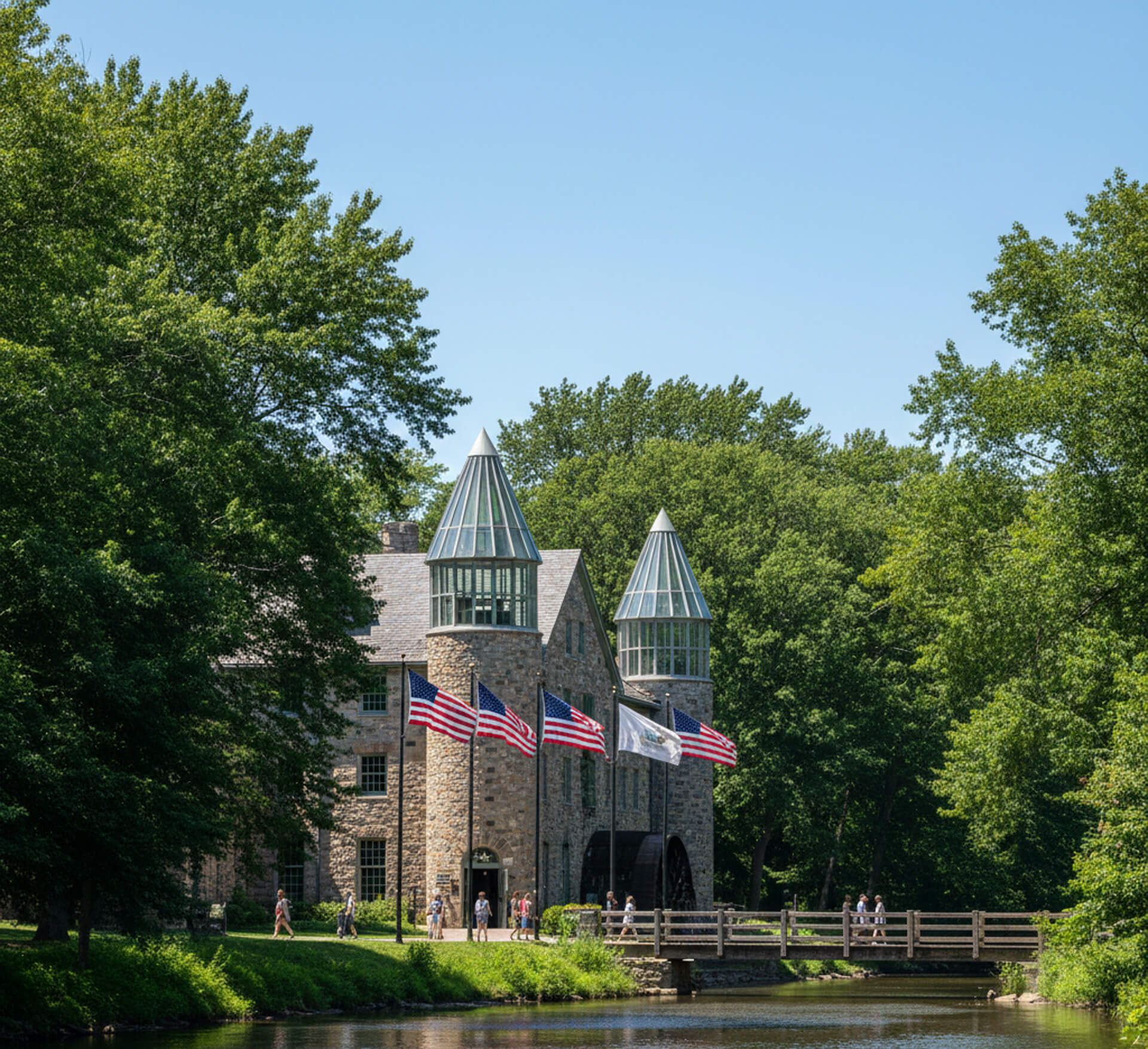 Historic stone building with glass peaks, surrounded by greenery, featuring American flags and people walking near a river in Chadds Ford, PA.