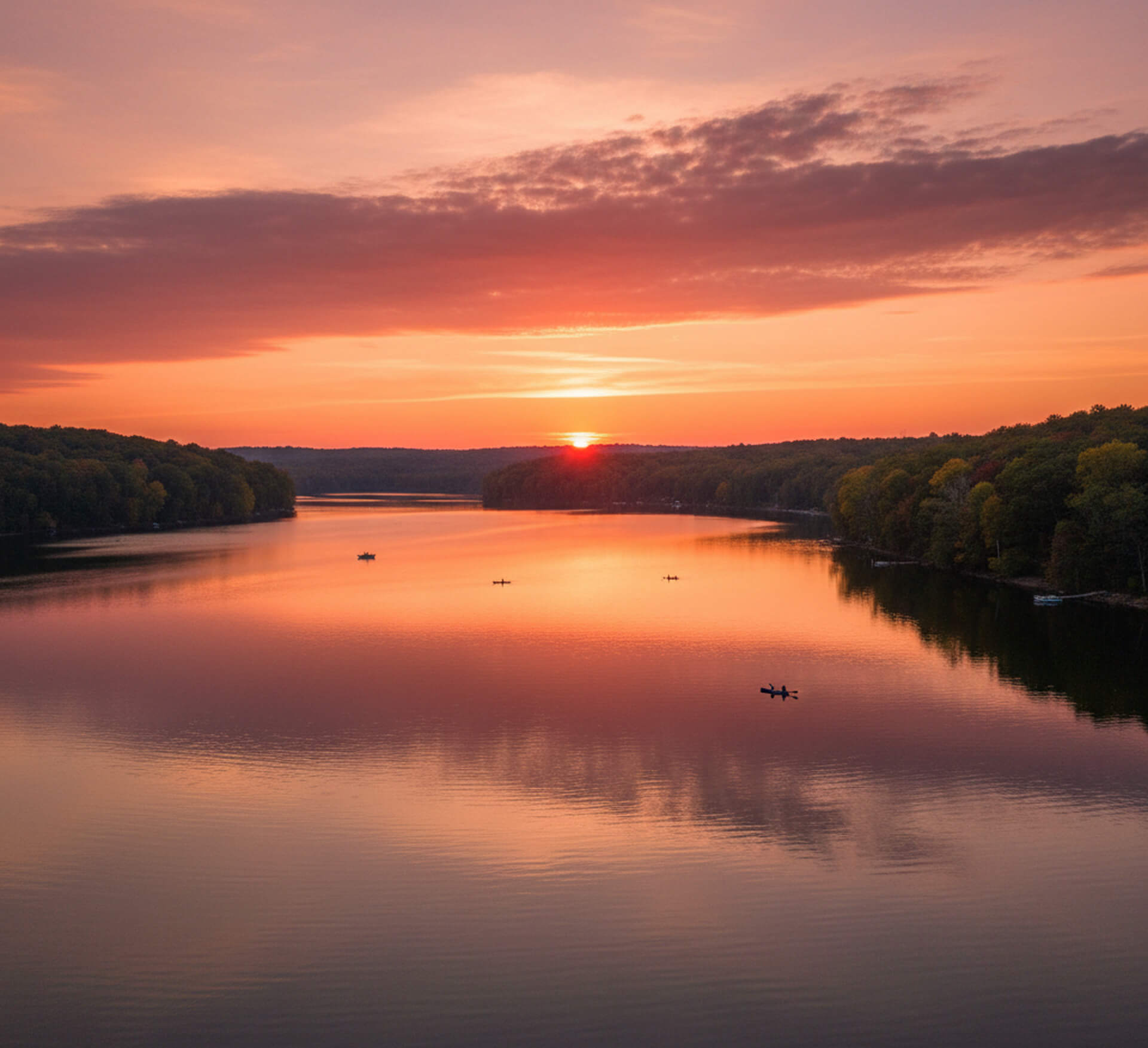Sunset over a tranquil lake in Exton, PA, with kayakers paddling on the water, surrounded by lush trees reflecting vibrant colors in the calm surface.